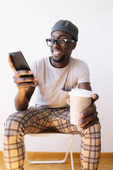 Smiling young man using mobile phone while sitting with coffee cup against white wall at home - JCMF00834