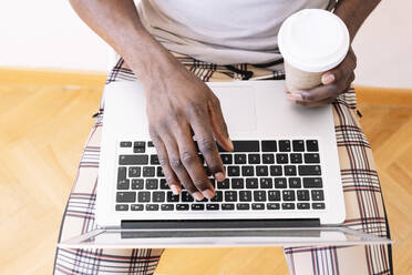 Junger Geschäftsmann, der einen Laptop benutzt, während er mit einer Kaffeetasse zu Hause sitzt - JCMF00835