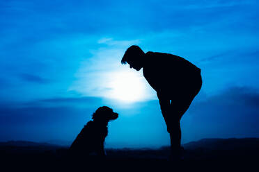 Silhouette eines jungen Mannes mit seinem Hund in blauem Nachtlicht - CAVF85051