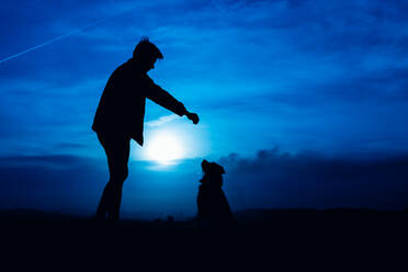 Silhouette eines jungen Mannes, der seinen Hund mit blauem Nachtlicht füttert - CAVF85052