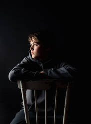 Low key portrait of adolescent boy sitting on a chair in a dark room. - CAVF85065