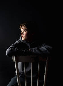 Low-Key-Porträt eines heranwachsenden Jungen, der auf einem Stuhl in einem dunklen Raum sitzt. - CAVF85065