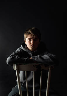 Low-Key-Porträt eines heranwachsenden Jungen, der auf einem Stuhl in einem dunklen Raum sitzt. - CAVF85066