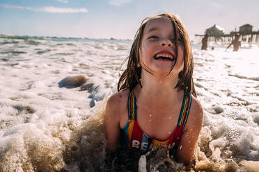 Young girl playing laying on beach while a wave crashes on her - CAVF85110