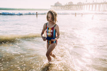 Young girl running through water at the beach with pier in background - CAVF85112