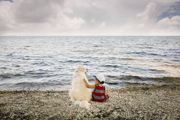Kleiner Junge sitzt mit Golden Retriever Hund am Strand und schaut auf den See - CAVF85156