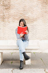 Lächelnde schöne Frau, die ein Buch liest, während sie auf einer Bank gegen die Wand sitzt - EYF05507