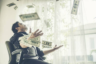 Successful Businessman Throwing Money While Sitting On Chair At Home - EYF05529