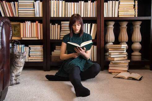 Junge Frau liest ein Buch, während sie gegen ein Bücherregal in der Bibliothek sitzt - EYF05645