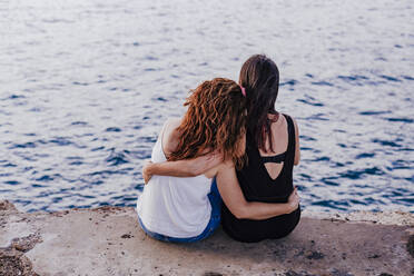 Back view of anonymous female friends in swimwear and summer clothes  embracing while standing on stony coast against waving sea during vacation  together in Puerto Escondido stock photo