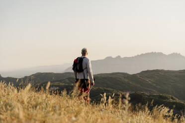 Aktiver älterer Mann mit Rucksack, der bei Sonnenuntergang auf eine Bergkette blickt - AFVF06683