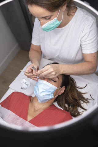 Mature female beautician using hand tools for eyelash extension on customer stock photo