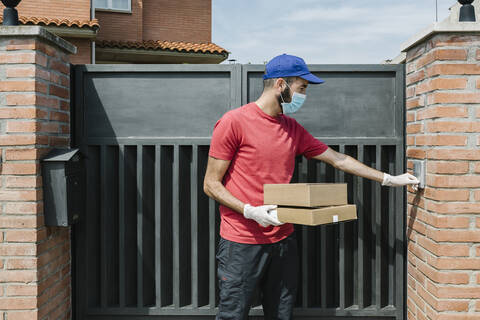 Male delivery person using intercom while standing with package at house gate during coronavirus stock photo