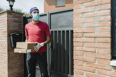 Mittlerer Erwachsener, männlicher Zusteller, der wegschaut, während er ein Paket in der Hand hält und vor dem Haustor steht - XLGF00188