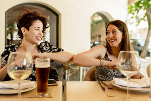 Smiling beautiful young women greeting with elbow bump while sitting at restaurant - EGAF00212
