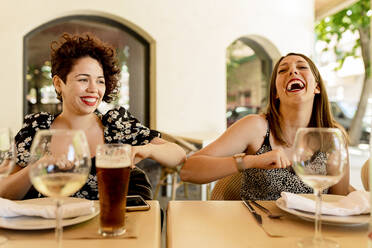 Fröhliche junge Frauen grüßen mit Ellbogenstoß beim Sitzen und Genießen im Restaurant - EGAF00213
