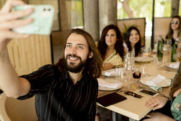 Lächelnder junger Mann, der ein Selfie mit weiblichen Freunden macht, die am Tisch im Restaurant sitzen - EGAF00217