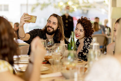 Lächelnder junger bärtiger Mann, der ein Selfie mit einer Frau macht, die an einem Tisch im Restaurant sitzt - EGAF00233