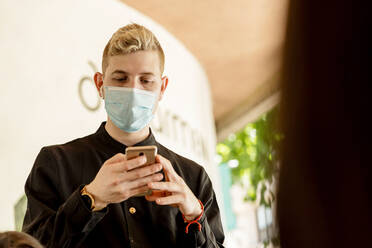 Young blond waiter with smart phone ordering while standing at restaurant - EGAF00235