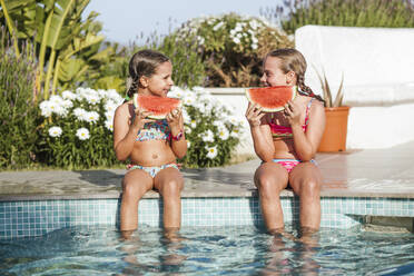 Lächelnde Zwillingsmädchen sitzen mit Wassermelonenscheibe am Pool an einem sonnigen Tag - LJF01573