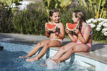 Happy twin girls sitting with watermelon slice at poolside on sunny day - LJF01574
