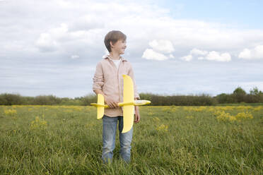 Smiling boy holding toy airplane while looking away on oilseed rape field against sky - EYAF01145