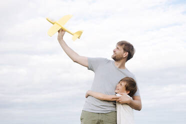 Glücklicher Junge, der seinen Vater umarmt und ein Spielzeugflugzeug gegen den Himmel hält - EYAF01148