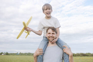 Lächelnder Mann mit Sohn auf den Schultern, der ein Spielzeugflugzeug gegen den Himmel hält - EYAF01150