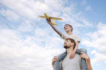 Man carrying son on shoulders playing with toy airplane against sky - EYAF01151