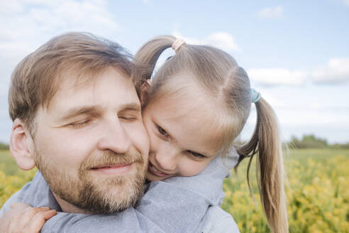 Lächelnder Mann mit geschlossenen Augen, der seine Tochter auf einem Feld gegen den Himmel huckepack nimmt - EYAF01154