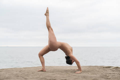 Flexible young naked woman practicing bending over backwards at beach against sky stock photo