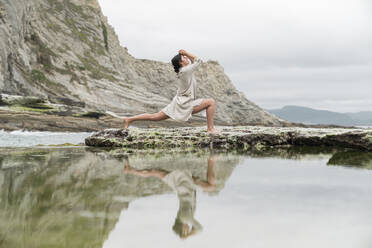 Reflection of young woman dancing on rock formation at beach - MTBF00443