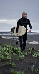 Junger Mann geht mit Surfbrett am Strand entlang in Island - CAVF85300