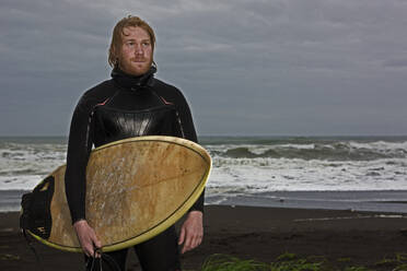 Junger Mann geht mit Surfbrett am Strand entlang in Island - CAVF85301