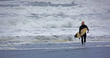 Junger Mann geht mit Surfbrett aus dem Wasser in Island - CAVF85302