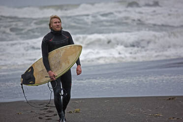 Junger Mann geht mit Surfbrett am Strand entlang in Island - CAVF85303