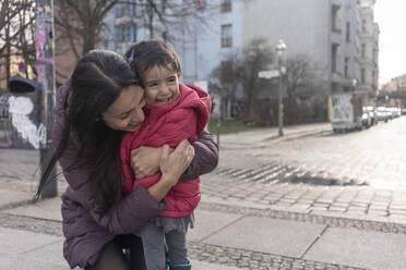 Glückliche Frau, die ihre Tochter umarmt, während sie auf einem Fußweg in der Stadt kniet - TAMF02285