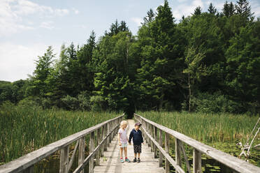 Brother and sister holding hands while standing on wooden footbridge over marsh - JVSF00009