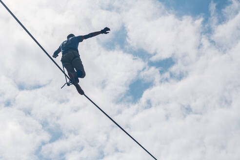 Low Angle View Of Man With Arms Outstretched Walking On Tightrope Against Cloudy Sky - EYF05964