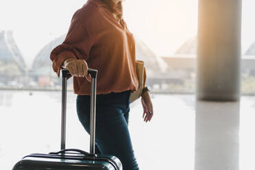 Midsection of Woman With Luggage Walking Against Window In Airport - EYF06491