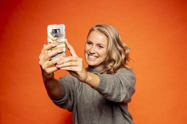 Glückliche blonde Frau nimmt Selfie gegen orange Hintergrund - DAWF01595