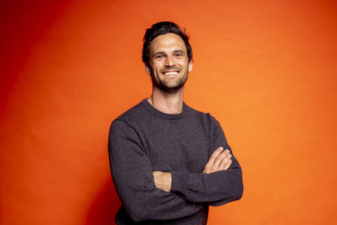 Smiling handsome man standing with arms crossed against orange background - DAWF01597
