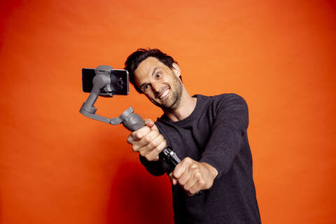 Cheerful man taking selfie while holding gimbal with smart phone against orange background stock photo