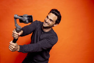 Playful gut aussehend Mann nimmt selfie durch Smartphone auf Gimbal gegen orange Hintergrund - DAWF01600