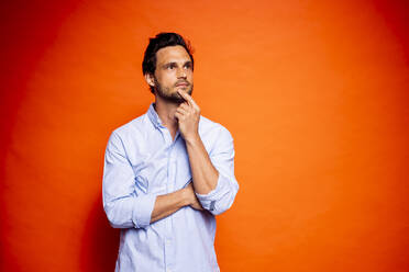 Thoughtful handsome man looking away while touching chin against orange background - DAWF01603