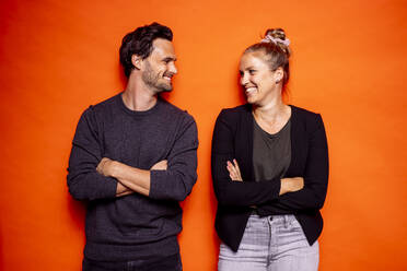 Glückliches erwachsenes Paar, das sich anschaut, während es mit verschränkten Armen vor einem orangefarbenen Hintergrund steht - DAWF01612