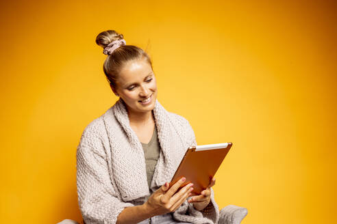 Lächelnde schöne Frau mit Haarknoten, die auf ein digitales Tablet schaut, während sie vor einem gelben Hintergrund sitzt - DAWF01613