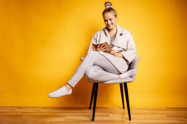 Smiling woman using digital tablet while sitting on chair against yellow background - DAWF01614