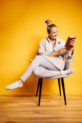 Happy woman using digital tablet while sitting on chair against yellow background - DAWF01615