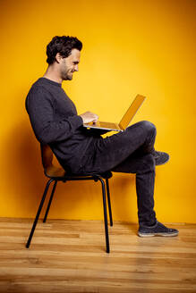 Smiling man using laptop while sitting on chair by yellow background - DAWF01616